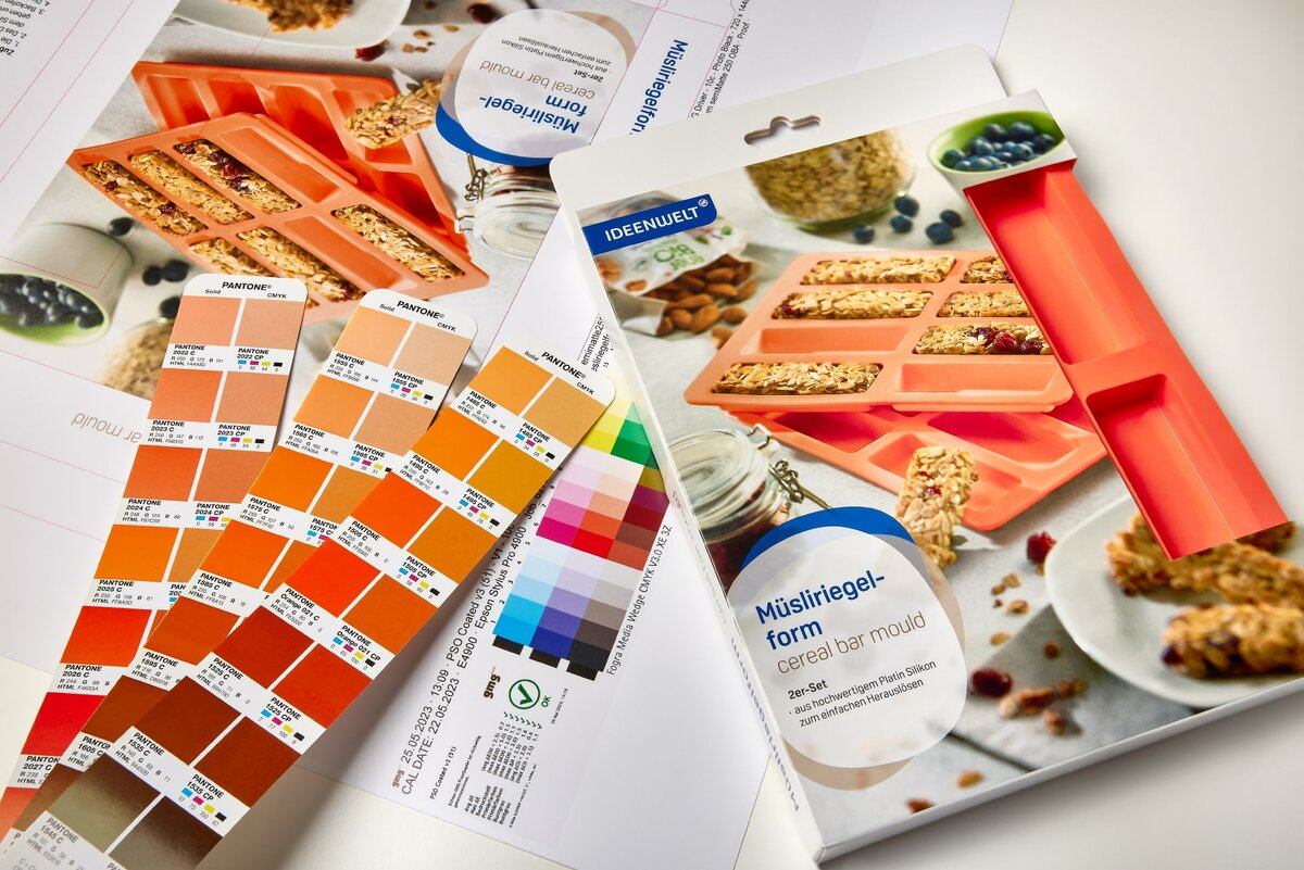 Das Foto zeigt die Verpackung einer orangefarbenen Müsliriegelform für die Rossmann Ideenwelt. Links daneben liegen Farbkarten aus einem Pantone-Fächer mit den vorgegebenen Tönen. Darunter ist die Stanze mit dem Color Code zu sehen.